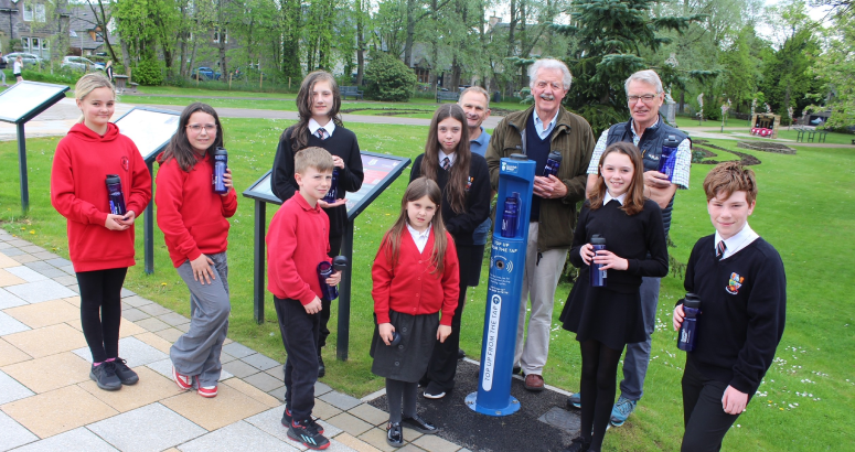 A group of children and adults stand outside. They are holding blue water bottles and surrounding a blue Scottish Water outdoor tap.