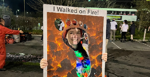 A woman stands smiling with a sign that reads "I Walked on Fire!".