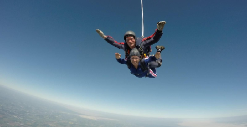 A man and woman smile as they skydive with clear blue skies behind them and a view of the land below.