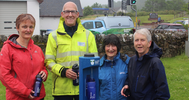A group of people are standing outside surrounding a blue Scottish Water branded water tap. They are holding blue plastic bottles and facing the camera. In the background is a white building with grey roof, and a Scottish Water branded vehicle.
