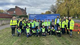 Students planting bulbs with GRP project team Group of people of various ages wearing safety vests, standing in front of a Scottish Water truck, in a suburban area.