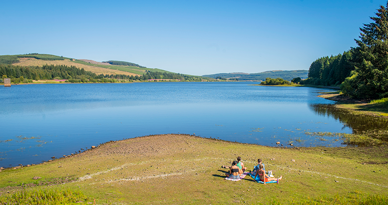 Carron Valley reservoir with group of people sunbathing on the shore on a sunny day